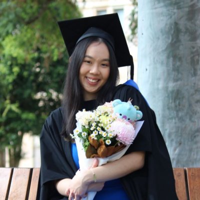 A young woman in graduation cap and gown sits on a bench seat in 澳门七星图's Great Court, clutching a bouquet of flowers with a small teddy bear inside it.
